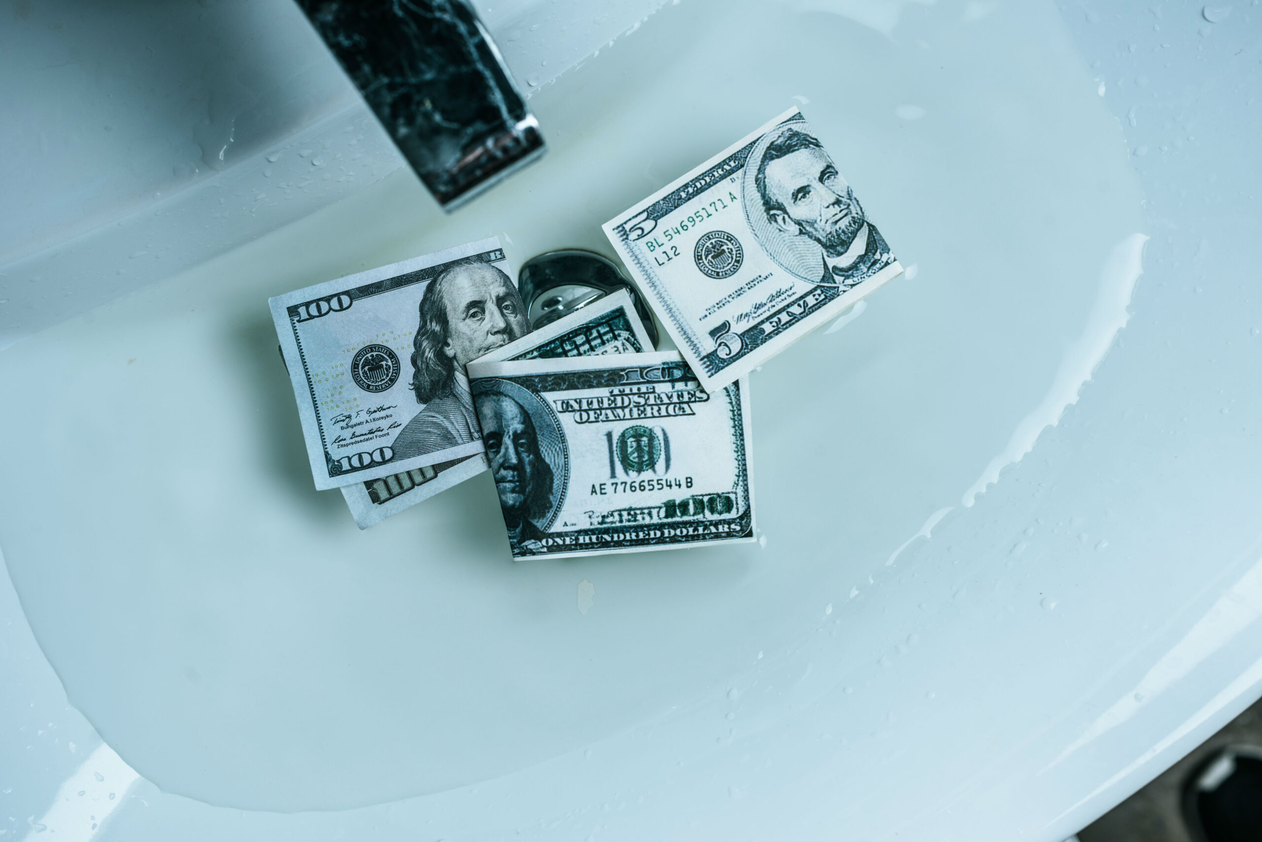 Several US dollar bills scattered in a white bathtub with a faucet nearby and water droplets on the tub surface.|informative image with money in a bath